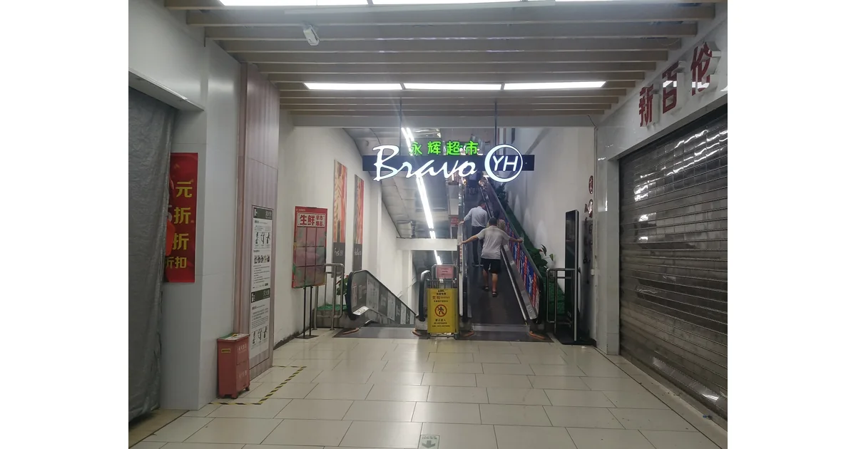 Entrada de supermercado com escadas rolantes, letreiro e clientes de costas.