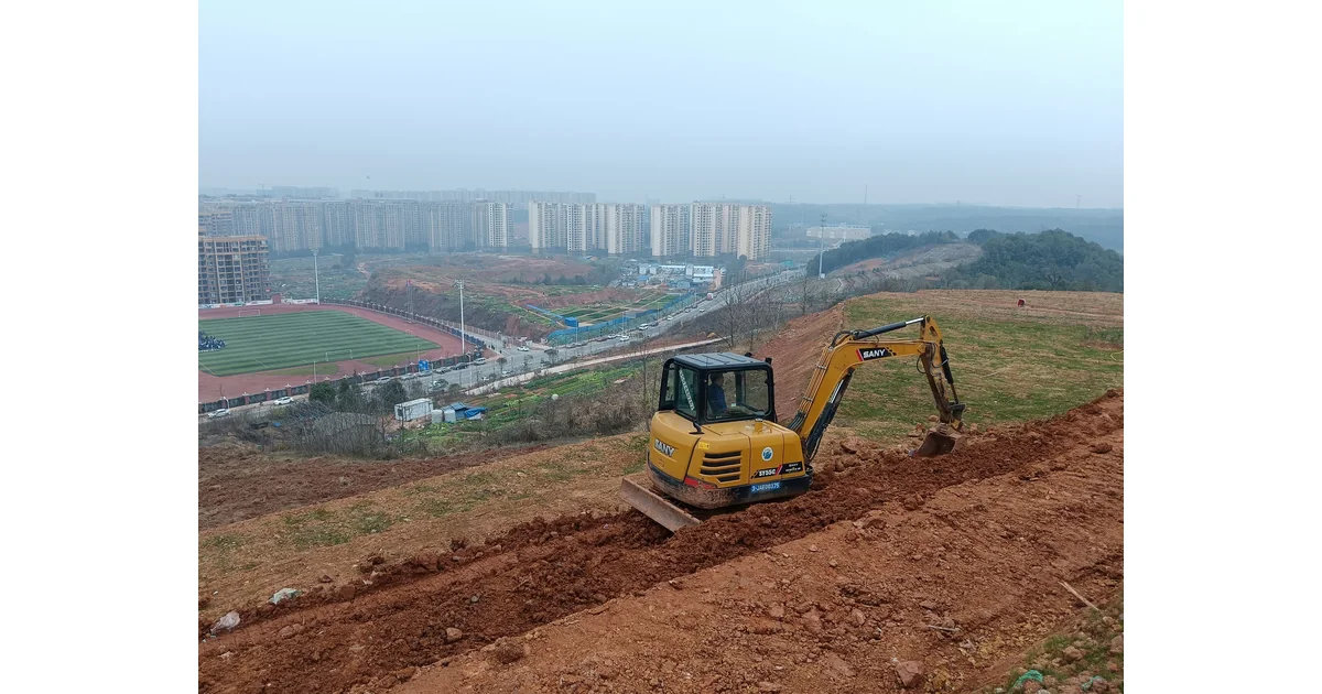 Escavadeira amarela em terreno de terra, com edifícios ao fundo.
